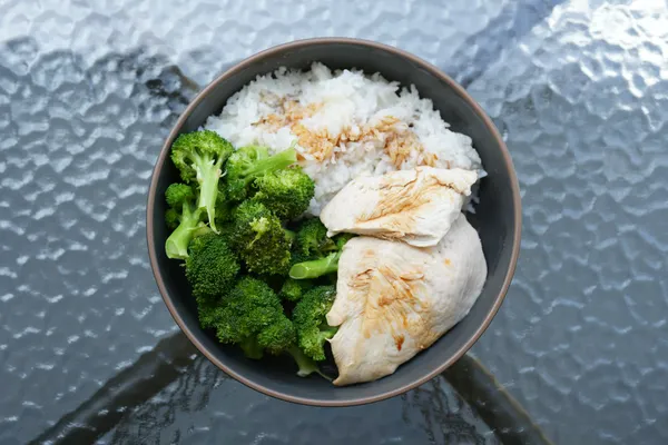 A bowl of chicken, rice and broccoli on a glass table